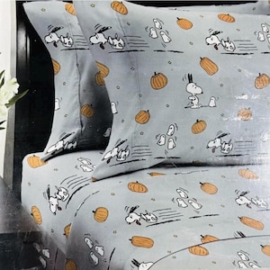 May include: A light gray bed sheet and pillowcase set featuring a repeating pattern of the cartoon character Snoopy running, orange pumpkins, and small white ghosts. The design includes black motion lines behind the character and small stars.