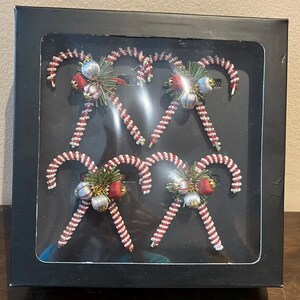 May include: A set of six candy cane ornaments in a black box. The candy canes are red and white, with a beaded design. Each candy cane has a cluster of red, silver, and green decorative elements.