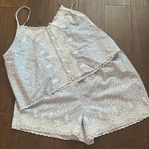 May include: A light blue two-piece set consisting of a camisole top and shorts. The top features thin straps, a button-down front, and white scalloped embroidery. The shorts have a matching white scalloped embroidered hem and a small floral print.