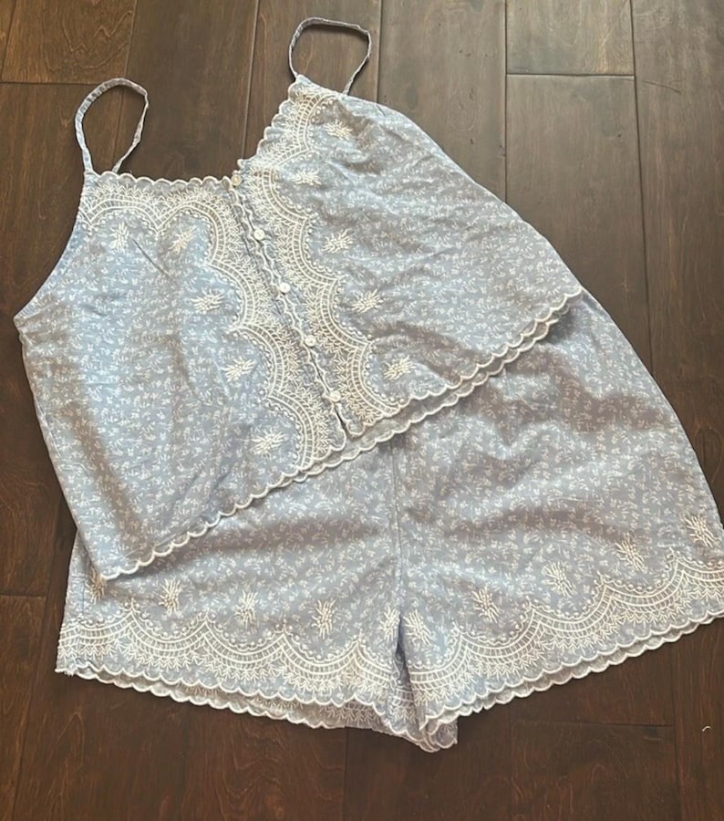 May include: A light blue two-piece set consisting of a camisole top and shorts. The top features thin straps, a button-down front, and white scalloped embroidery. The shorts have a matching white scalloped embroidered hem and a small floral print.