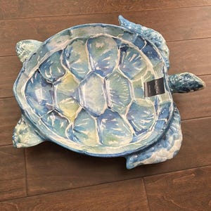 May include: A decorative serving dish in the shape of a sea turtle. The dish is painted in shades of blue and green, with a watercolor effect. The dish has a label that says "NAUTICA".