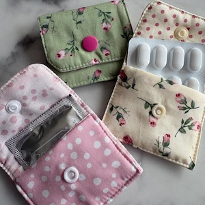 May include: Three small fabric pouches with snap closures. One is green with pink roses and a pink snap. One is cream with pink roses and a white snap, holding a blister pack of pills. One is pink with white dots and a white snap, holding a foil packet.