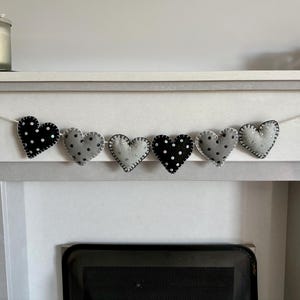 Könnte beinhalten: Eine handgefertigte Herzgirlande mit sechs Filzherzen. Die Herzen wechseln sich ab zwischen grau mit weißen Punkten und schwarz mit weißen Punkten. Jedes Herz ist mit einem Kontrastfaden genäht und auf einer weißen Schnur aufgereiht. Die Girlande wird auf einem weißen Kaminsims präsentiert.