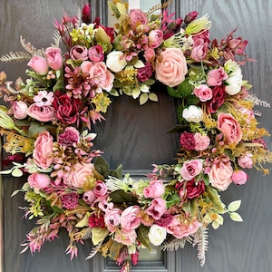 May include: A circular floral wreath featuring a mix of pink, white, and burgundy flowers and foliage. The wreath is displayed on a grey door. The arrangement includes roses, peonies, and other blooms, creating a dense and textured display.