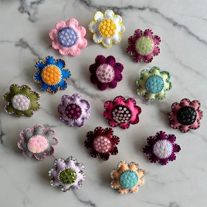Puede incluir: Surtido de broches de flores de fieltro en varios colores y patrones. Cada broche presenta una base de fieltro de color diferente con un centro lleno de lentejuelas o cuentas.