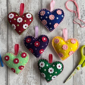 May include: Six felt heart ornaments with different colored felt and flower embellishments. The hearts are hanging from a white wooden surface.