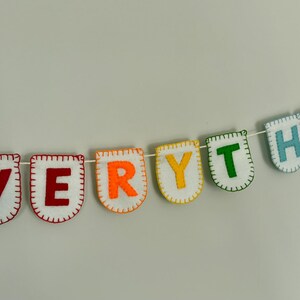 Può includere: Un colorato striscione in feltro con la scritta "EVERYTHING" in singoli stendardi con lettere. Ogni stendardo è bianco con un bordo colorato e una lettera di colore diverso. Lo striscione è infilato su uno spago bianco con pompon gialli ad ogni estremità.