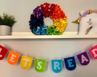 Puede incluir: Una colorida corona de fieltro con un patrón de arcoíris cuelga sobre una estantería. A la derecha, una cabeza de unicornio blanco con cuerno dorado y melena rosa. Una pancarta debajo dice "LET'S READ" en letras de fieltro.