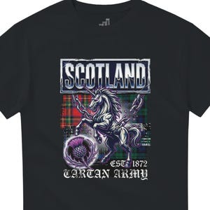 Puede incluir: Camiseta negra con un diseño gráfico que presenta la palabra "SCOTLAND" en un marco blanco. El diseño incluye un unicornio, un cardo y el texto "EST. 1872 TARTAN ARMY". El fondo tiene un patrón de tartán.