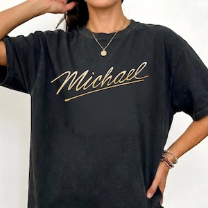May include: Black oversized t-shirt featuring the name "Michael" in gold script. The shirt has a vintage, washed-out look. This t-shirt is a casual, comfortable style.