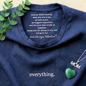 Könnte beinhalten: Ein marineblaues T-Shirt mit dem Wort "everything" am unteren Rand. Das Shirt hat Text in der Nähe des Kragens, der lautet: "because that's exactly what you are to me..." Eine silberne "MOM"-Halskette und ein grüner herzförmiger Stein sind ebenfalls sichtbar.