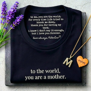 May include: A navy blue t-shirt with white text that reads "to the world, you are a mother." A gold necklace with an "M" pendant and a wooden heart pendant that says "mom" are on the shirt. Lavender sprigs are on the left.