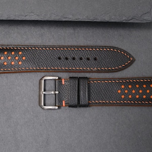 May include: Black leather watch strap with orange stitching and perforated details. The strap has a silver-tone buckle. The design includes a series of small, evenly spaced holes. The strap is designed for a classic timepiece.