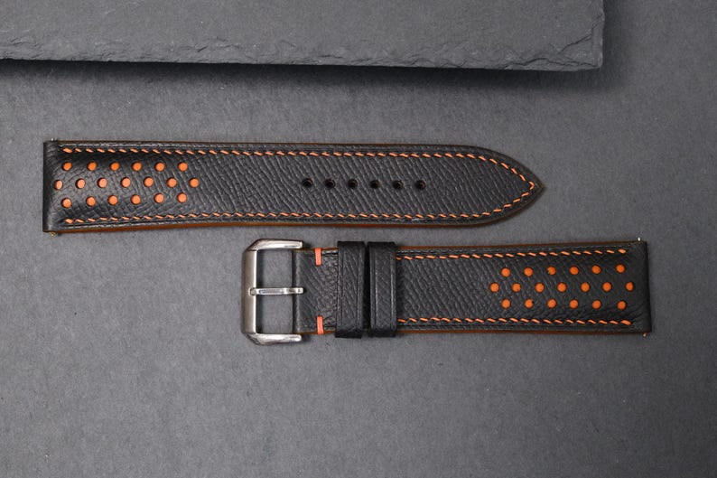 May include: Black leather watch strap with orange stitching and perforated details. The strap has a silver-tone buckle. The design includes a series of small, evenly spaced holes. The strap is designed for a classic timepiece.