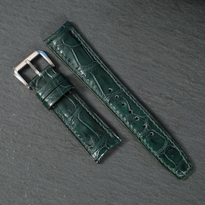 Könnte beinhalten: Ein dunkelwaldgrünes Uhrenarmband mit einer glänzenden, geprägten Alligator-Textur. Das Armband verfügt über eine polierte silberfarbene Metallschnalle und eine Reihe gestanzter Löcher zur Anpassung, was ein raffiniertes Design zeigt.