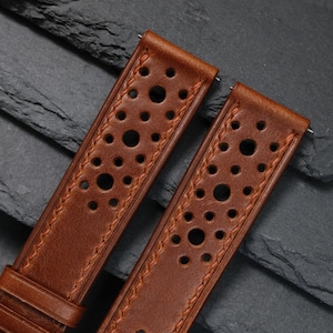 K&ouml;nnte beinhalten: Zwei braune Leder-Uhrenarmb&auml;nder mit perforiertem Design. Die Armb&auml;nder haben einen gen&auml;hten Rand und sind vor einem dunklen, strukturierten Hintergrund platziert. Die Armb&auml;nder sind zur Befestigung an einer Uhr konzipiert.