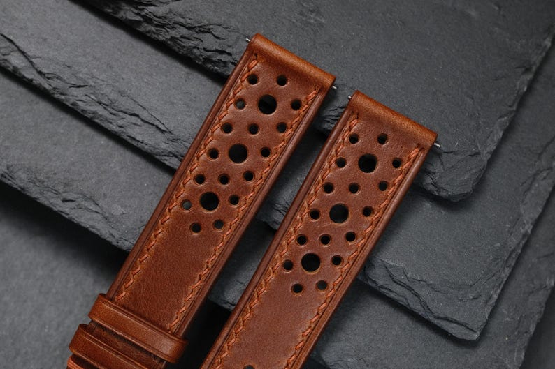K&ouml;nnte beinhalten: Zwei braune Leder-Uhrenarmb&auml;nder mit perforiertem Design. Die Armb&auml;nder haben einen gen&auml;hten Rand und sind vor einem dunklen, strukturierten Hintergrund platziert. Die Armb&auml;nder sind zur Befestigung an einer Uhr konzipiert.