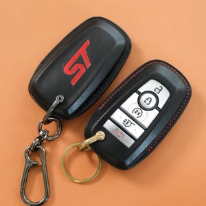 Op de afbeelding: Twee zwarte lederen autosleutelhoezen met rode stiksels. Eén hoes heeft een rood ST-logo en een donker metalen karabijnhaak. De andere hoes toont de zilveren knoppen van een autosleutel, inclusief iconen voor vergrendelen, ontgrendelen en starten.