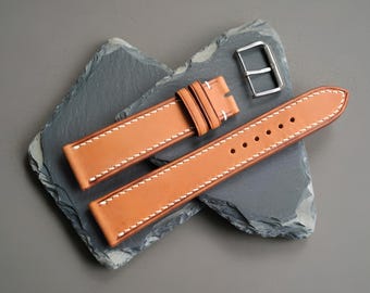 Könnte beinhalten: Ein beiges Leder-Uhrenarmband mit weißen Kontrastnähten an den Kanten, zwei Lederschlaufen und einer separaten silberfarbenen Metall-Dornschließe. Das Armband besteht aus zwei Teilen mit einem spitz zulaufenden Ende und mehreren Löchern.