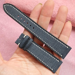 May include: A pair of dark blue watch straps with white stitching. The straps are made of a textured material and have a classic design. The watch straps are held in a hand against a pink background.