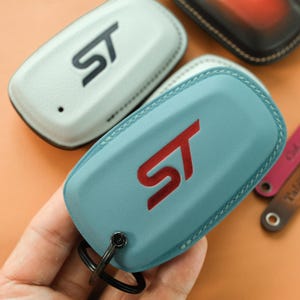 May include: A light blue leather key fob cover featuring a red embossed ST logo on the front and a black metal ring attachment. The cover has visible stitching along the edges and a smooth, rounded rectangular shape for a car key fob.