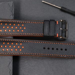 May include: Black leather watch strap with orange stitching and perforated detailing. The strap has a stainless steel buckle and orange underside. A small screwdriver is also present.