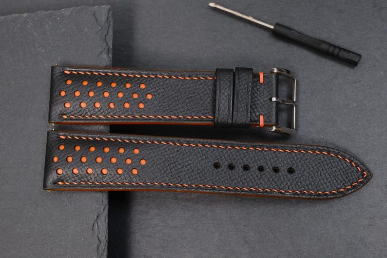 May include: Black leather watch strap with orange stitching and perforated detailing. The strap has a stainless steel buckle and orange underside. A small screwdriver is also present.