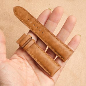 Może przedstawiać: Para jasnobrązowych skórzanych pasków do zegarków. Paski są trzymane w dłoni i wykonane są z teksturowanego materiału skórzanego. Paski mają przeszycia wzdłuż krawędzi i otwory na sprzączkę zegarka.
