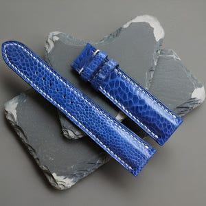 May include: Two blue watch straps with white stitching, made from a textured material, possibly leather. The straps are displayed on a gray surface with dark gray, irregularly shaped stones.
