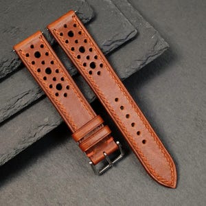 Può includere: Due cinturini per orologi in pelle marrone con un design traforato e cuciture a contrasto. Un cinturino ha una fibbia argentata. I cinturini sono angolati su una superficie grigio scuro, mostrando la consistenza e i dettagli della pelle.