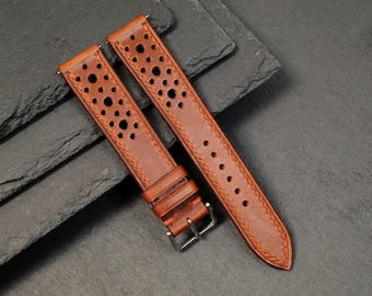 May include: Two brown leather watch straps with a perforated design and contrasting stitching. One strap has a silver buckle. The straps are angled on a dark gray surface, showcasing the texture and detail of the leather.