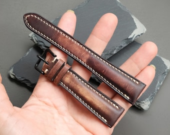 Könnte beinhalten: Ein zweiteiliges Uhrenarmband aus braunem Leder mit Used-Optik, cremefarbenen Kontrastnähten an den Rändern und einer mattschwarzen Metallschnalle. Das Armband hat ein zulaufendes Design mit sichtbarer Maserung und einem dunklen Farbton.