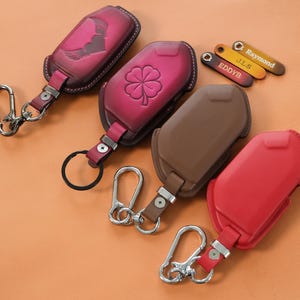 May include: Four leather car key fob covers with metal keychains. One is dark pink with a heart and wings, one is pink with a four-leaf clover, one is brown, and one is red. Three small leather tags are labeled EDDYB, J.L.S, and Raymond.