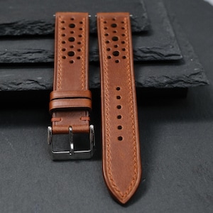 K&ouml;nnte beinhalten: Zwei braune Leder-Uhrenarmb&auml;nder mit perforiertem Design. Die Armb&auml;nder haben eine Edelstahlschnalle und sind vor einem dunklen Hintergrund platziert. Die Armb&auml;nder sind neu und ungetragen.
