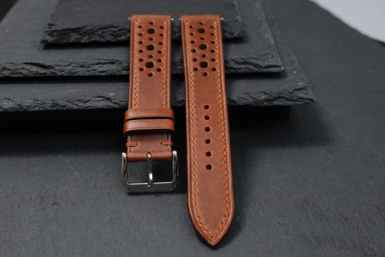 K&ouml;nnte beinhalten: Zwei braune Leder-Uhrenarmb&auml;nder mit perforiertem Design. Die Armb&auml;nder haben eine Edelstahlschnalle und sind vor einem dunklen Hintergrund platziert. Die Armb&auml;nder sind neu und ungetragen.