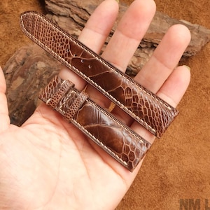 Puede incluir: Un par de correas de reloj marrones con un patrón texturizado similar a la piel de reptil. Las correas son de cuero y tienen costuras blancas. La imagen está sobre un fondo marrón.