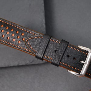 May include: Black leather watch strap with orange stitching and perforated details. The strap has a stainless steel buckle. The watch band is designed for a classic and sporty look. The strap's measurements are in centimetres.