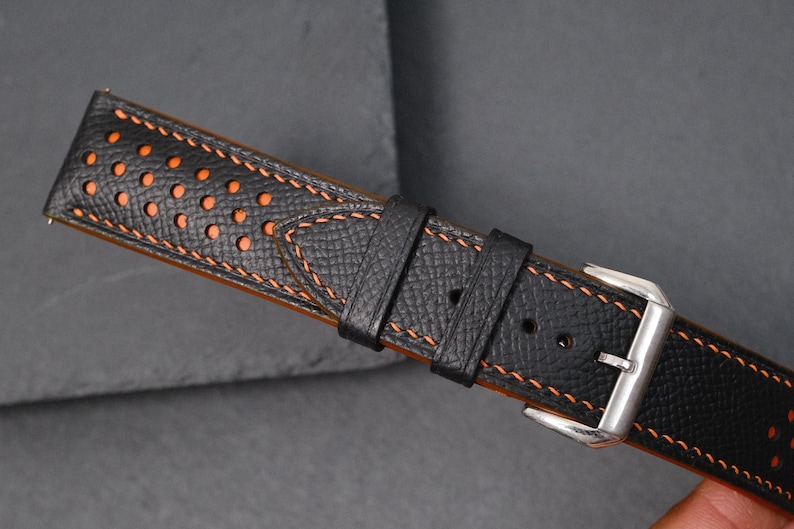 May include: Black leather watch strap with orange stitching and perforated details. The strap has a stainless steel buckle. The watch band is designed for a classic and sporty look. The strap's measurements are in centimetres.