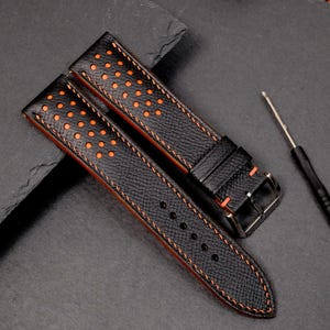 May include: Black leather watch strap with orange stitching and orange dot perforations. The strap has a black buckle and a small screwdriver. The strap is shown on a dark grey surface.