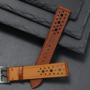 K&ouml;nnte beinhalten: Zwei braune Leder-Armb&auml;nder mit perforiertem Design. Eines der Armb&auml;nder ist heller als das andere. Die Armb&auml;nder liegen auf einem dunkelgrauen Hintergrund, mit einer silbernen Schnalle an einem Armband.