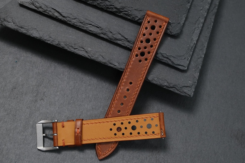 K&ouml;nnte beinhalten: Zwei braune Leder-Armb&auml;nder mit perforiertem Design. Eines der Armb&auml;nder ist heller als das andere. Die Armb&auml;nder liegen auf einem dunkelgrauen Hintergrund, mit einer silbernen Schnalle an einem Armband.