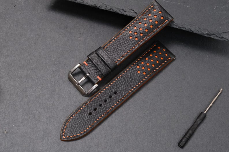 May include: Black watch strap with orange stitching and perforated holes. The strap has a stainless steel buckle and a matching black keeper. A small screwdriver is also included.