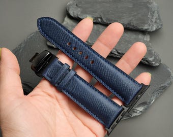 May include: A navy blue watch strap with a textured surface and a black metal deployment clasp. The strap features matching blue stitching along the edges and includes black metal connectors designed to attach to a smartwatch device.