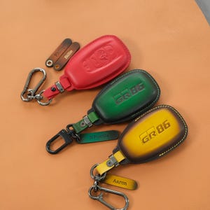 Könnte beinhalten: Drei Autoschlüsselhüllen aus Leder in Rot, Grün und Gelb mit dunklen Rändern. Die grünen und gelben Hüllen tragen den geprägten Schriftzug GR86. Jede Hülle enthält einen Metallkarabiner und einen kleinen, mit Namen personalisierten Lederanhänger.