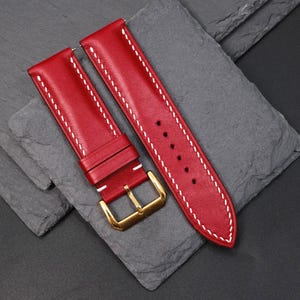 May include: Red leather watch strap with white stitching and a gold-coloured buckle. The strap is shown on a dark grey surface. The watch band is new and unused.