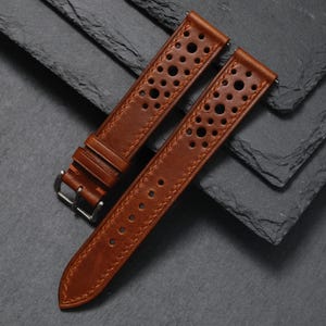 K&ouml;nnte beinhalten: Zwei braune Leder-Uhrenarmb&auml;nder mit perforiertem Design und silberner Schnalle. Die Armb&auml;nder sind aus reichhaltigem, strukturiertem Leder gefertigt und f&uuml;r eine klassische Uhr konzipiert. Die Armb&auml;nder werden auf einer dunkelgrauen Oberfl&auml;che pr&auml;sentiert.