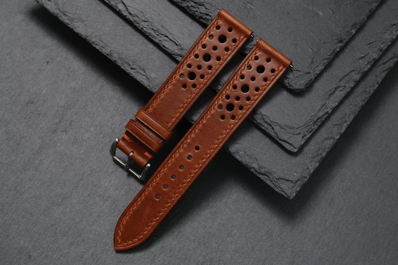 K&ouml;nnte beinhalten: Zwei braune Leder-Uhrenarmb&auml;nder mit perforiertem Design und silberner Schnalle. Die Armb&auml;nder sind aus reichhaltigem, strukturiertem Leder gefertigt und f&uuml;r eine klassische Uhr konzipiert. Die Armb&auml;nder werden auf einer dunkelgrauen Oberfl&auml;che pr&auml;sentiert.