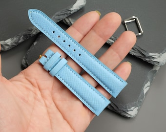 May include: A light blue watch strap with a textured surface and matching stitching. The strap consists of two pieces, one with a series of adjustment holes and the other with a fixed loop and a floating loop, accompanied by a separate metal buckle.
