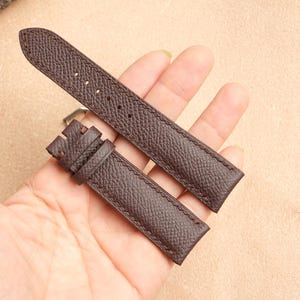 May include: A pair of dark brown leather watch straps with a textured surface. The straps are held in a hand, showcasing their color and design. The watch straps are a classic accessory.