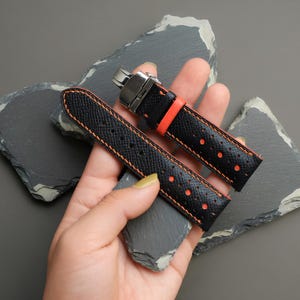 May include: Black watch straps with orange stitching and accents. The straps have a textured surface and are perforated with small holes. A silver clasp is attached to one end. The straps are displayed on a grey background.